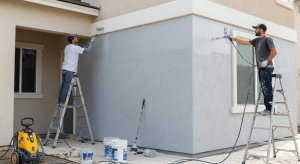 Workers painting a stucco exterior wall of a house using ladders and paint sprayers
